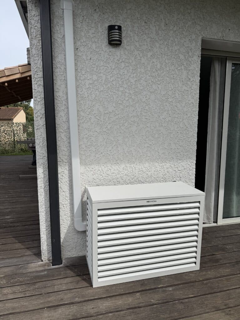 Cachage d'unité extérieure de climatisation, modèle DECO'Clim, de couleur grise, posé sur une terrasse en bois sombre près d'un mur texturé et d'une gouttière.