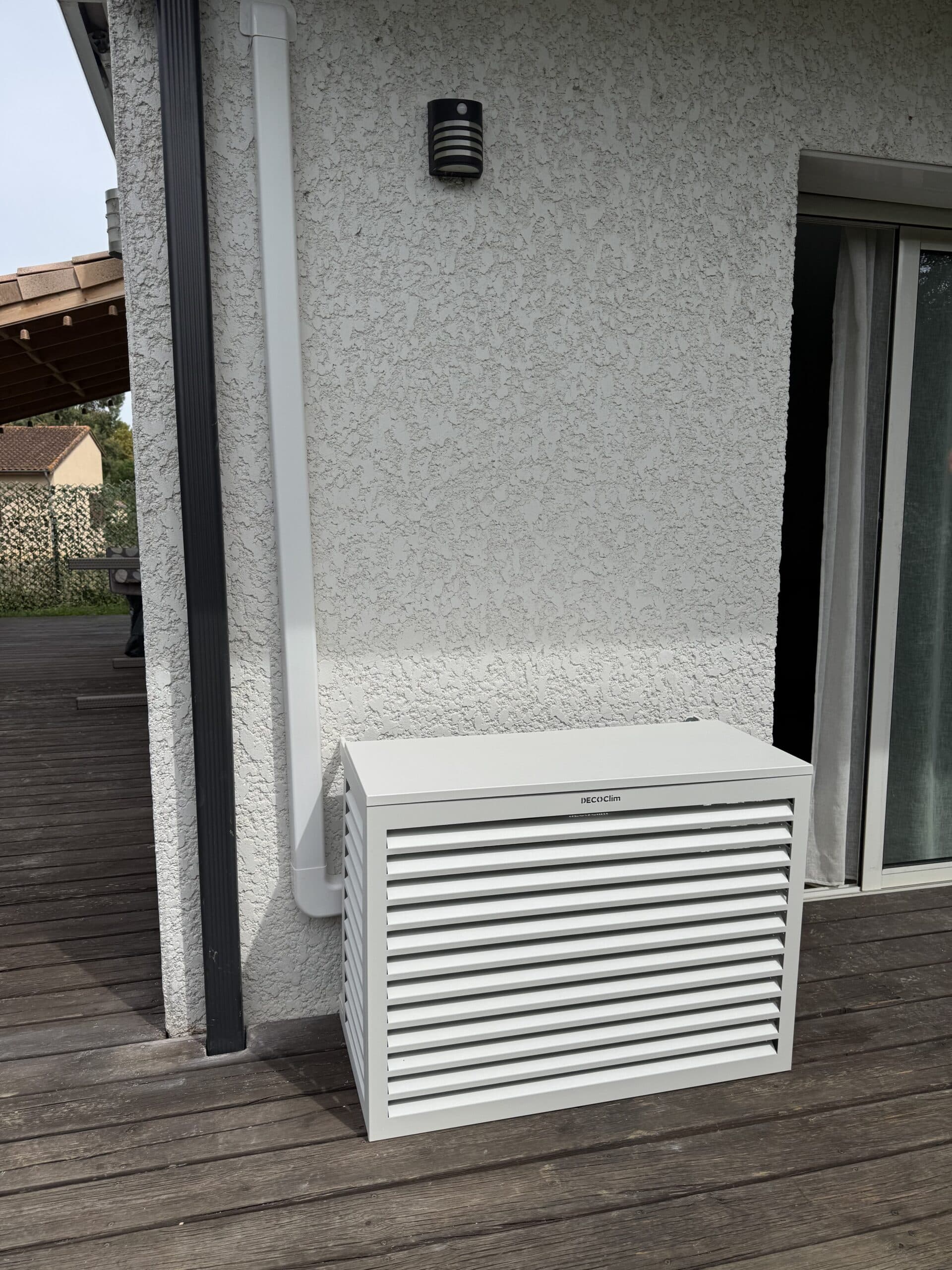 Cachage d'unité extérieure de climatisation, modèle DECO'Clim, de couleur grise, posé sur une terrasse en bois sombre près d'un mur texturé et d'une gouttière.