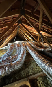 Intérieur d'un grenier avec charpente en bois, isolation et plusieurs conduits de ventilation flexibles et brillants (gainés) convergeant vers le centre sous les chevrons.