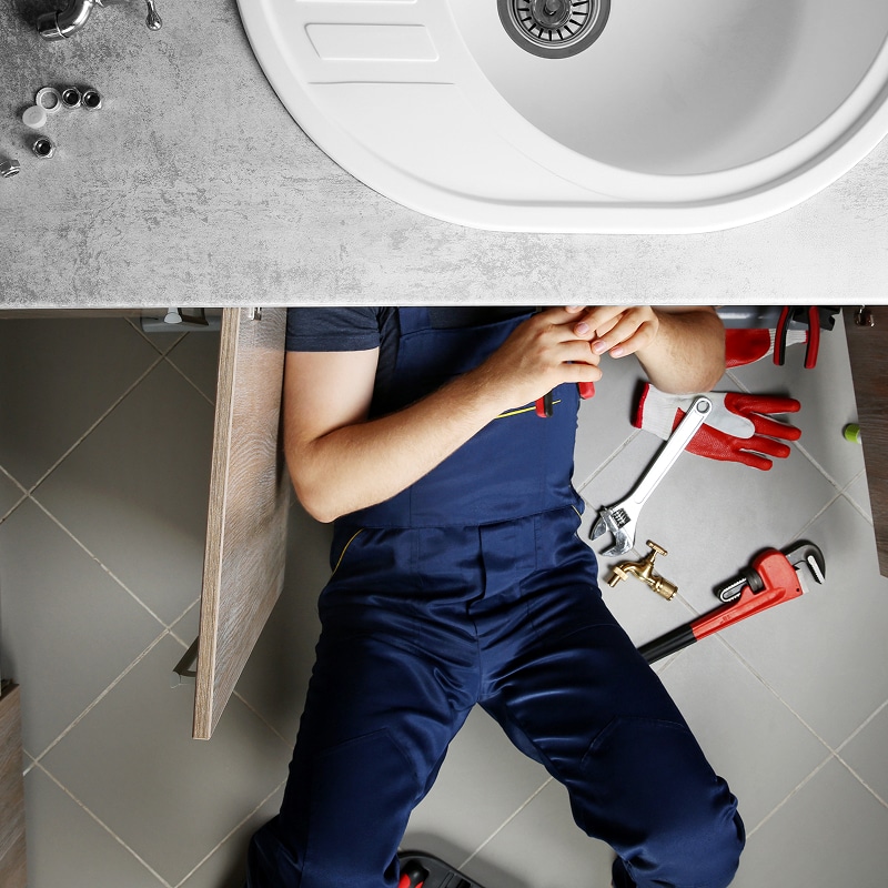 Plombier en salopette bleue réparant sous un évier blanc, outils et robinet sur le sol carrelé gris.