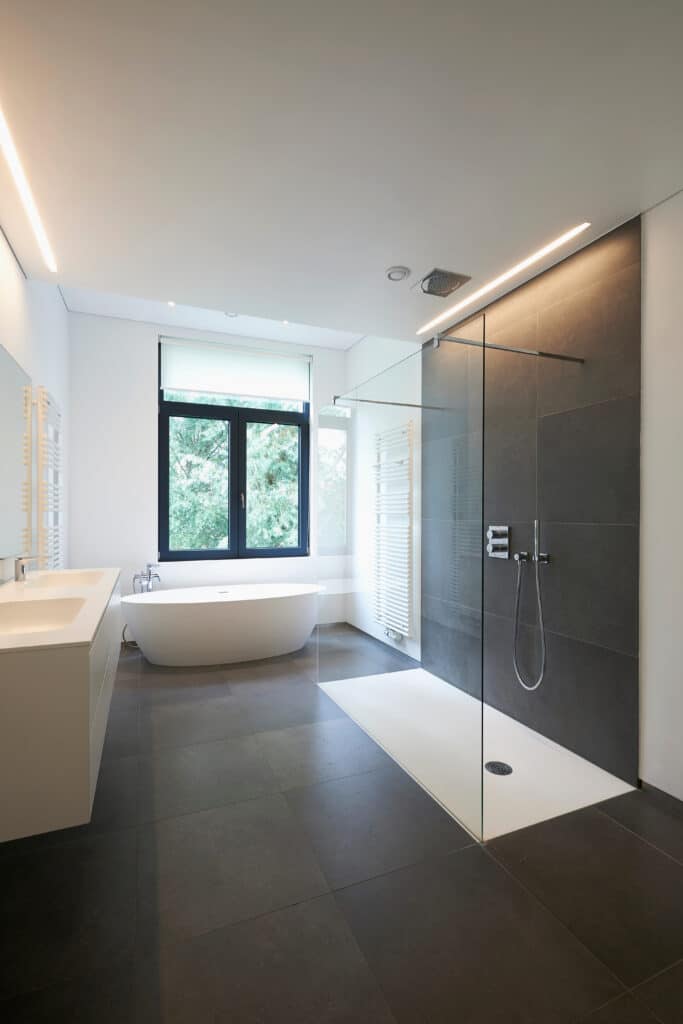Salle de bain moderne avec grande douche carrelée gris foncé, baignoire îlot blanche, double vasque et fenêtre sur la verdure.