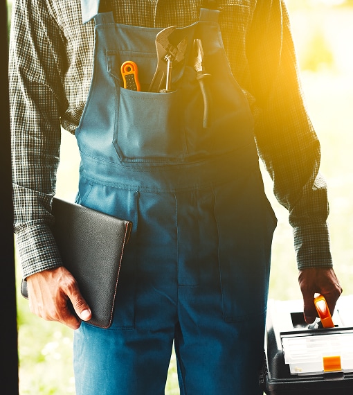 Technicien en salopette bleue tenant une tablette et une boîte à outils, prêt à intervenir sous un éclairage solaire intense.