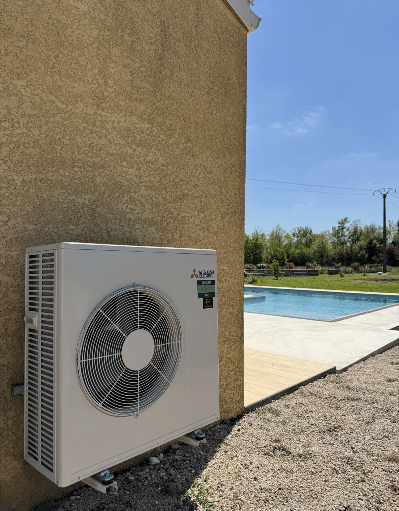 Unité extérieure de pompe à chaleur Mitsubishi Electric Mr.Slim Inverter R32 installée près d'une maison à crépi jaune, avec une piscine visible en arrière-plan.